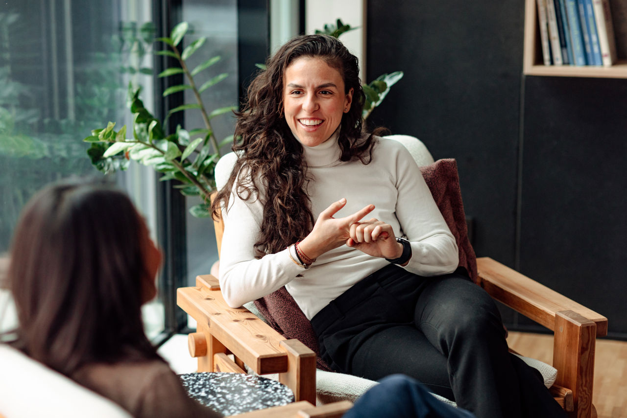 Woman sitting in chair in office talking, other woman seen from back listening